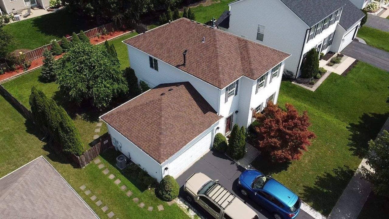 Aerial view of a recently installed warm brown architectural asphalt shingle roof on a two-story white suburban house with an attached garage. The new roof shows crisp lines, precise flashing around the chimney, and clean ridge caps. The home is surrounded by green lawns and mature landscaping, reflecting a complete exterior renovation and premium roof upgrade.